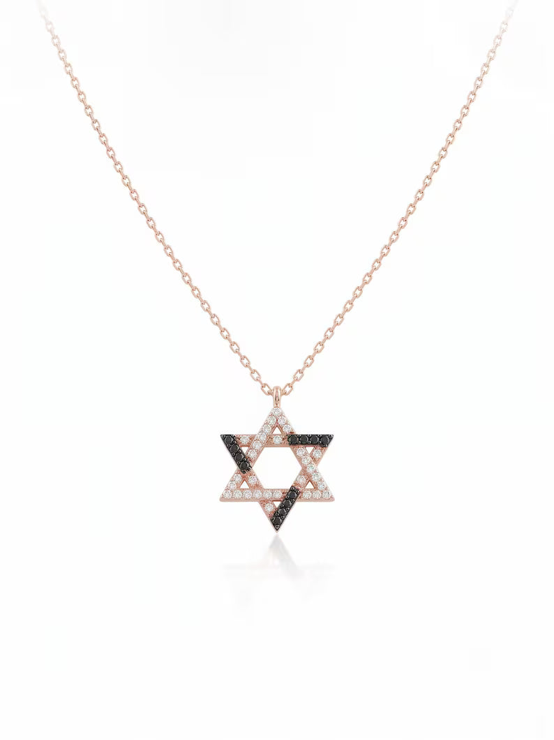 A delicate rose gold chain necklace featuring a Star of David pendant. The pendant is embellished with small white stones on two sides and dark black stones on the other, set against a clean white background with a subtle reflection.