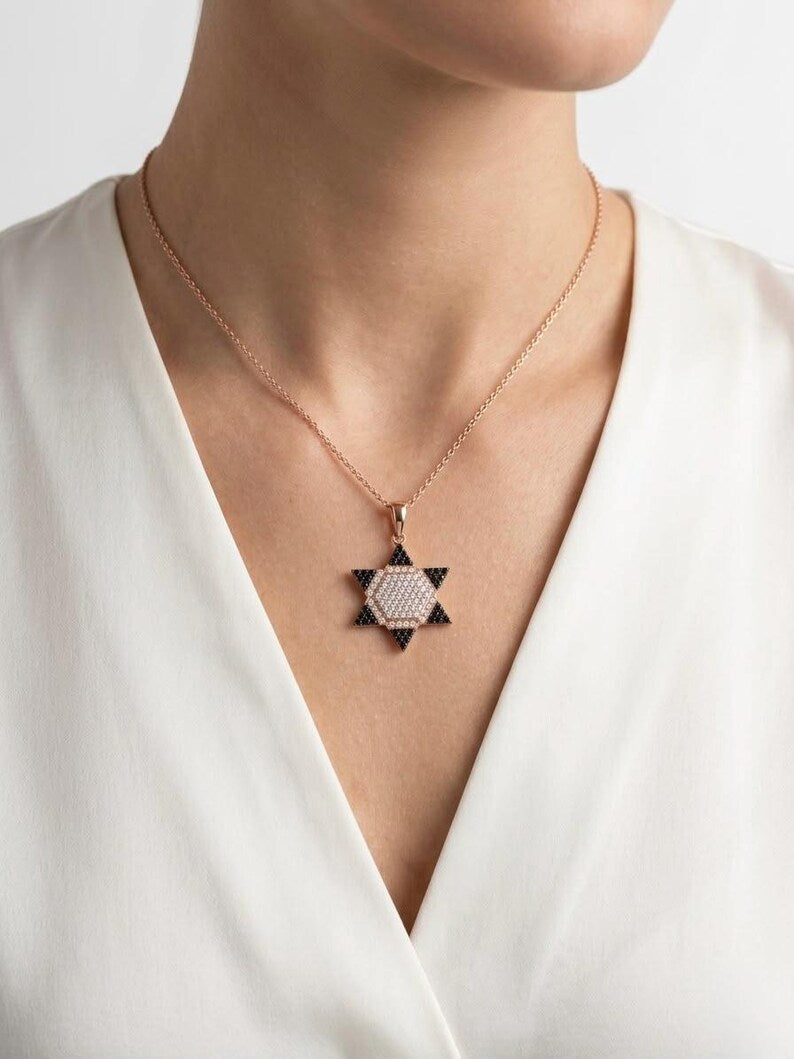 A close-up of a model wearing a delicate rose gold chain necklace with a geometric star pendant. The pendant features a textured light center with dark triangular accents, styled against a white V-neck top on a clean white background.