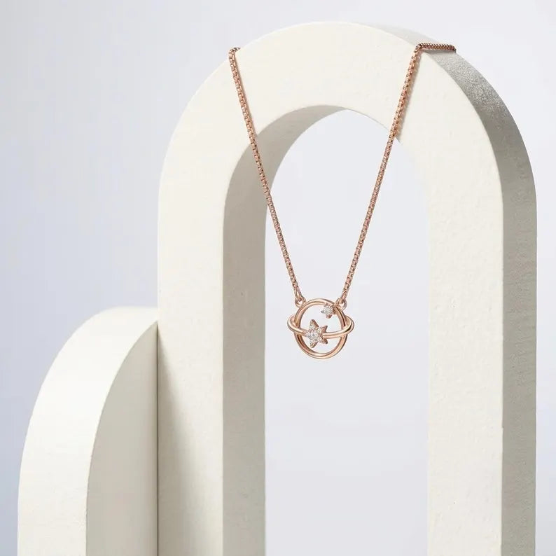 A rose gold necklace with a double-ring pendant and a small star detail, displayed on a white geometric stand against a light gray background.