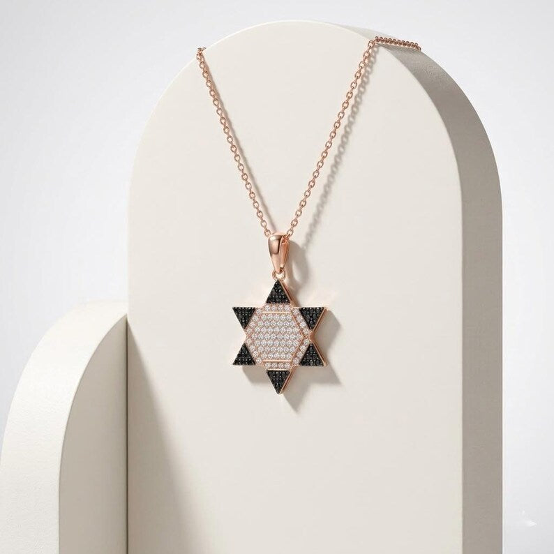 A rose gold Star of David pendant necklace with a black and white patterned center, displayed on a cream-colored arched stand.
