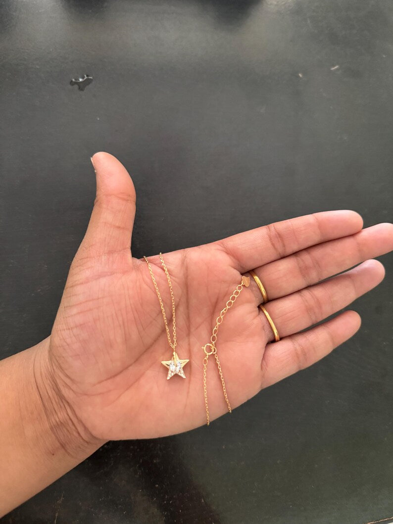A gold necklace with a star-shaped pendant and an adjustable chain extension, held in a person's open palm against a dark gray background.