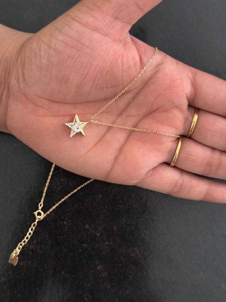 A gold star pendant necklace with a clear central stone, draped across an open hand. The hand also has two gold rings on the fingers, and the background is a dark, textured surface.
