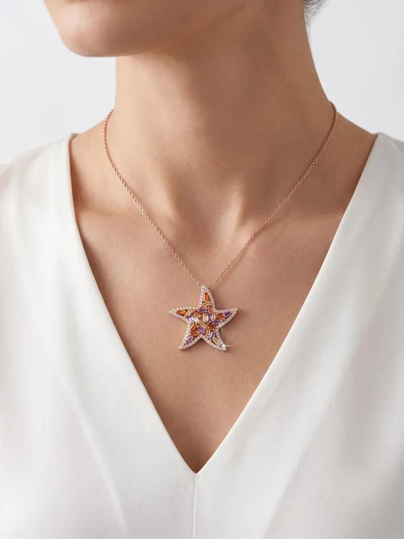 A close-up of a model wearing a rose gold starfish pendant necklace with small orange and white stones, styled with a white V-neck top.
