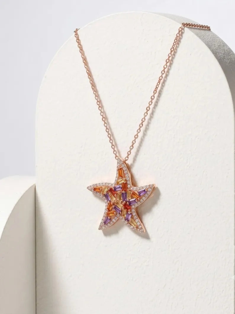 A rose gold starfish pendant necklace with orange and purple gemstone accents, displayed on a white textured stand.