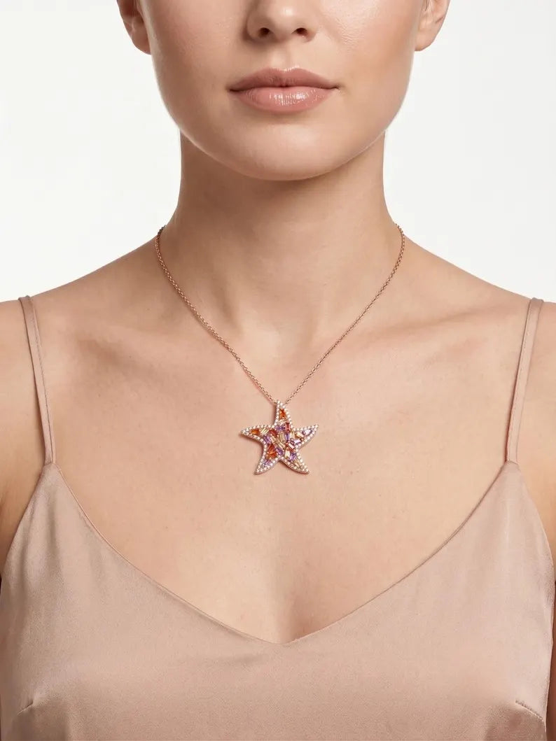 A close-up of a model wearing a rose gold starfish pendant necklace with pink and orange gemstone accents, styled with a beige satin camisole top against a clean white background.