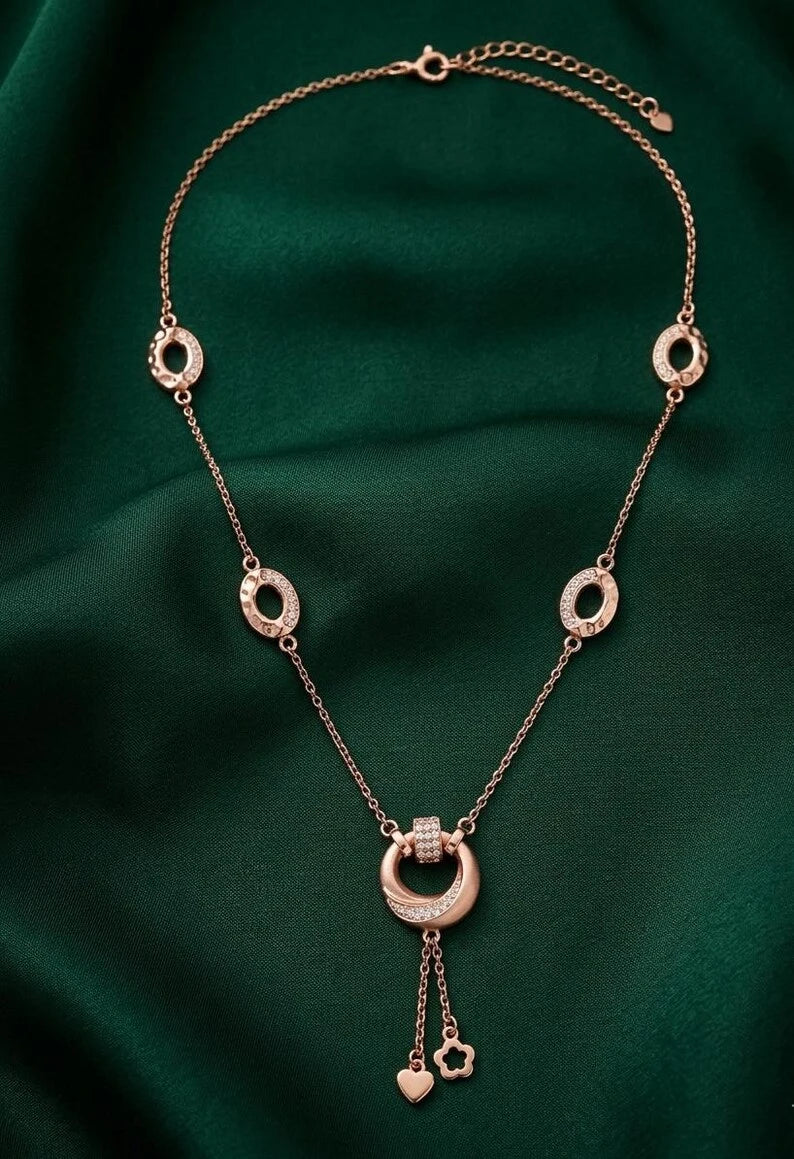A rose gold necklace with circular and crescent moon motifs, displayed on a draped dark green satin fabric background.