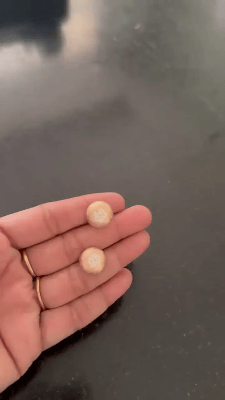 Two small round beige jewelry pieces held in a person's hand against a dark reflective surface.