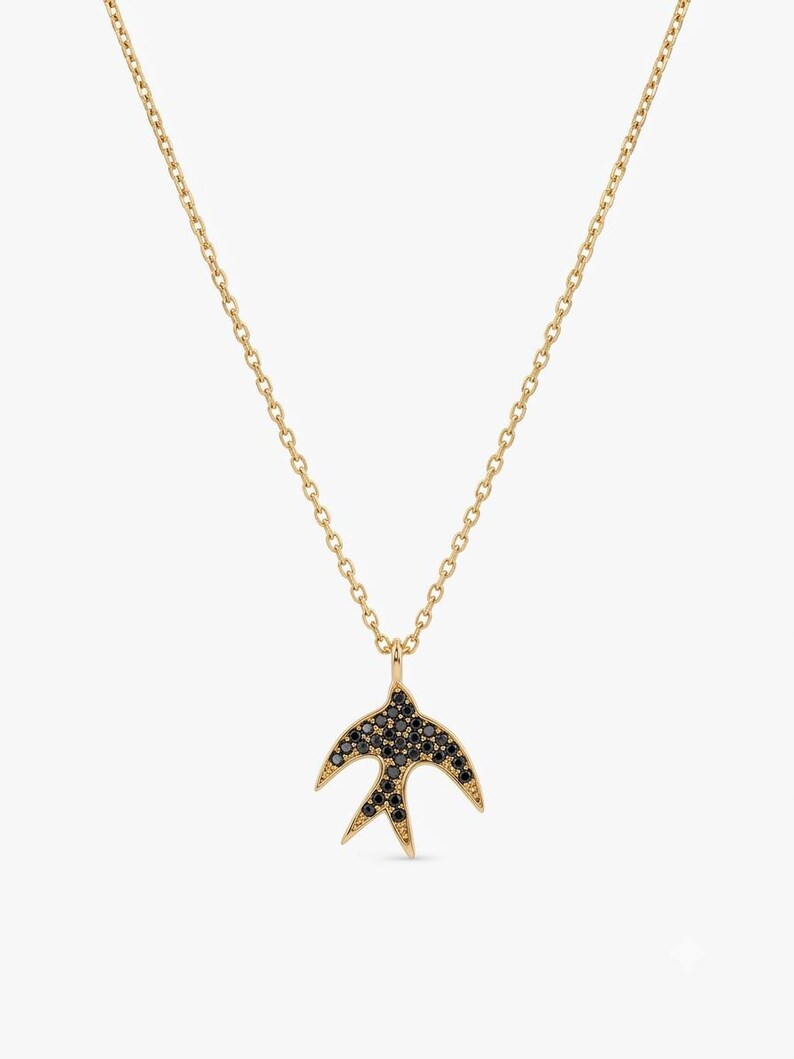 A gold chain necklace featuring a pendant shaped like a swallow, embellished with small dark stones, set against a plain light gray background.