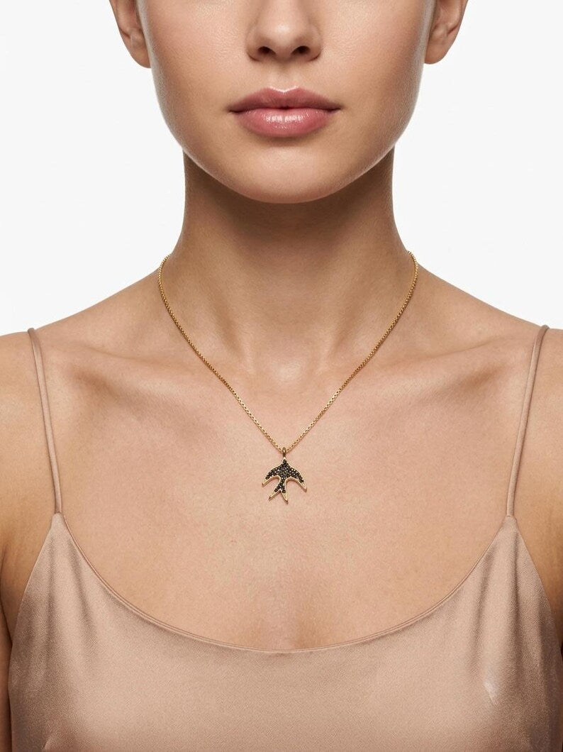 A close-up of a model wearing a delicate gold chain necklace with a small bird pendant, styled with a beige camisole top against a clean white background.