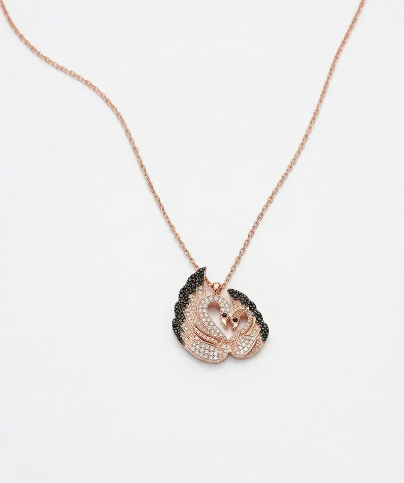 A rose gold necklace with a pendant shaped like two embracing swans, one wing covered in small black stones and the other in clear rhinestones, on a plain white background.