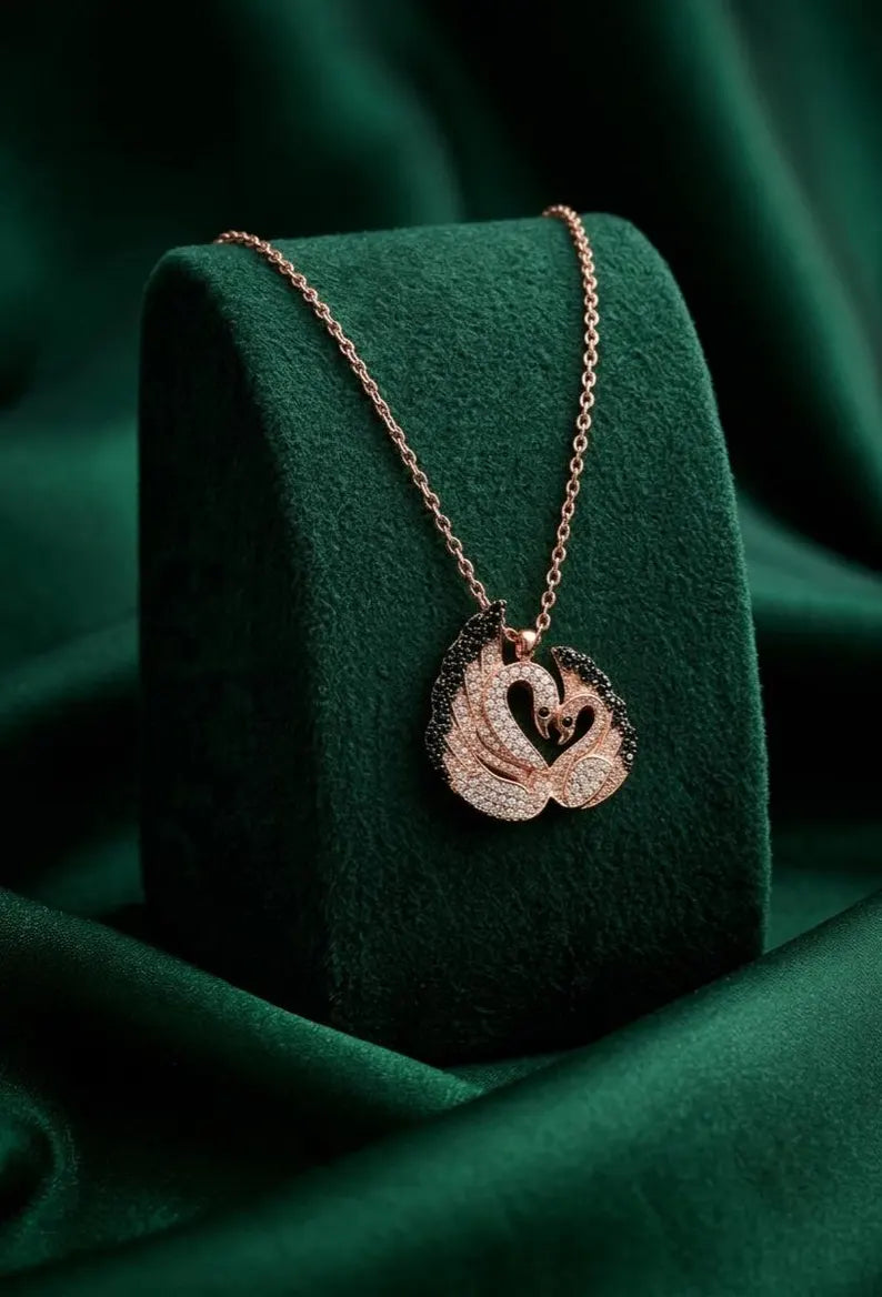 A rose gold necklace with a swan pendant displayed on a dark green velvet jewelry stand against a draped green fabric background.