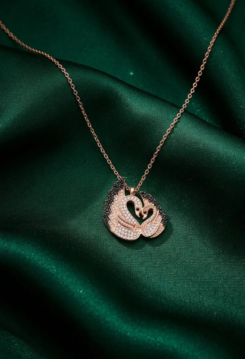 A rose gold necklace with a swan pendant, displayed on a draped dark green satin fabric background.