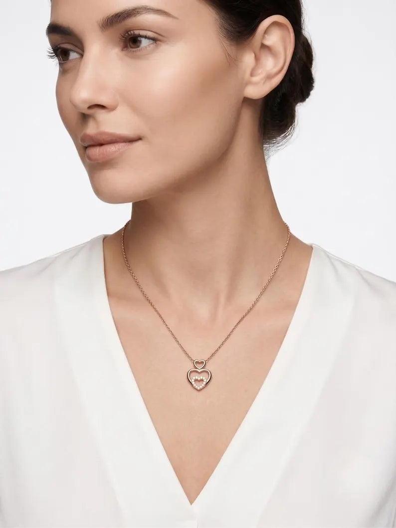 A close-up of a woman wearing a delicate rose gold heart pendant necklace with a small heart on a larger heart, styled with a white V-neck top against a light gray background.