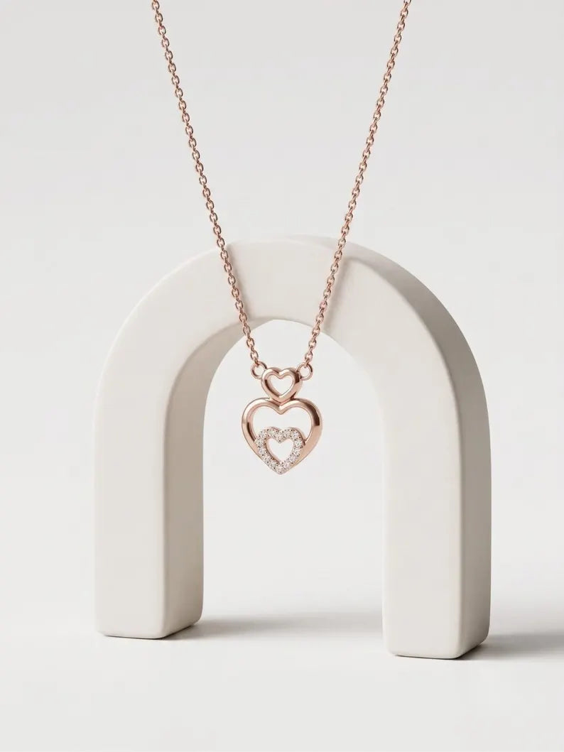 A rose gold heart pendant necklace with a smaller inner heart paved with clear stones, hanging from a delicate chain and displayed on a white arch stand against a light gray background.