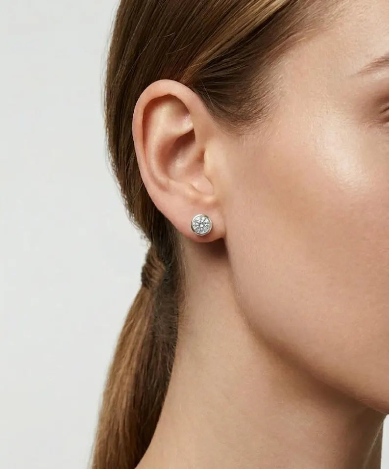 A close-up of a woman's ear wearing a small round stud earring with a silver-toned floral or starburst setting and clear stones, against a light neutral background.