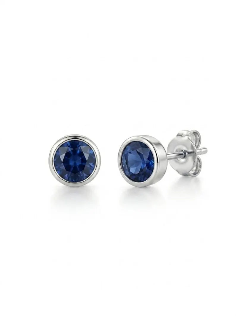 A pair of round bezel-set stud earrings featuring deep blue faceted gemstones in polished silver-toned metal, with one earring shown from the front and the other from the side/back.