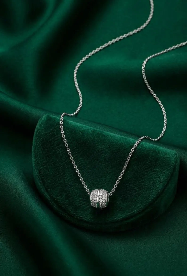 A silver chain necklace with a small round pavé pendant resting on a dark green velvet pouch, set against a matching dark green satin fabric background.