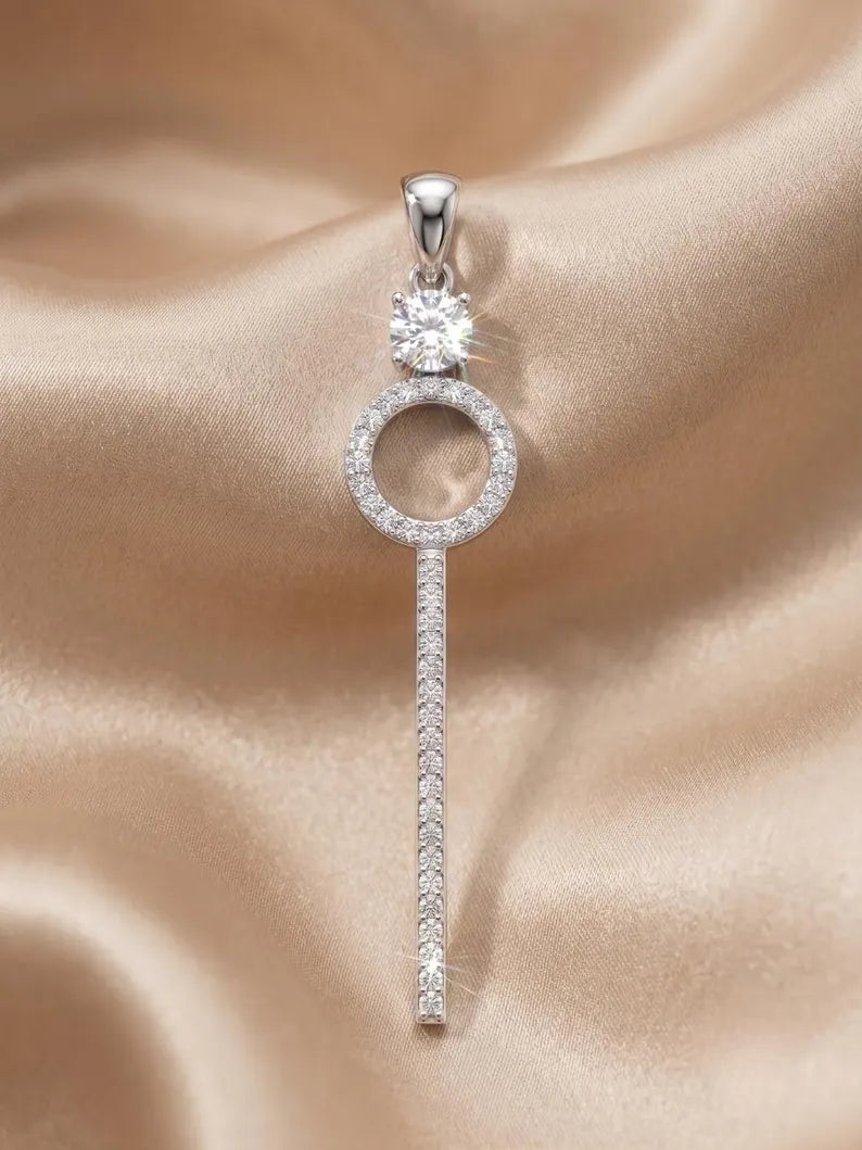 A silver key-shaped pendant with a round clear gemstone at the top and a vertical bar paved with small clear stones, displayed on a beige satin fabric background.
