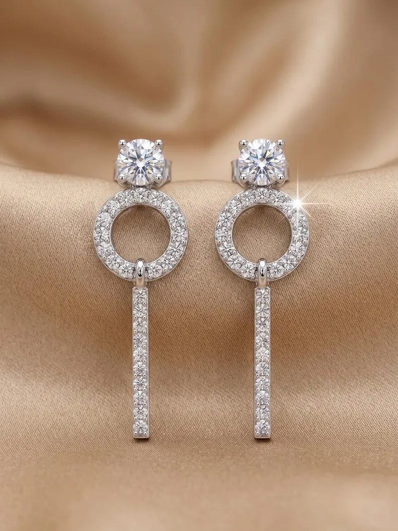 A pair of silver-toned drop earrings with round bezel-set clear stones, circular pavé rings, and vertical pavé bars, displayed on a draped beige satin fabric.