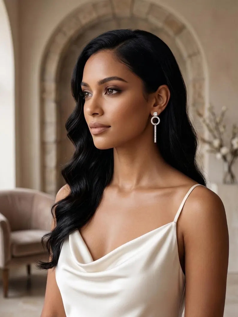 A woman with long, wavy black hair wearing a cream satin slip dress and a long silver key-shaped dangle earring, posed in front of a warm beige stone archway background.