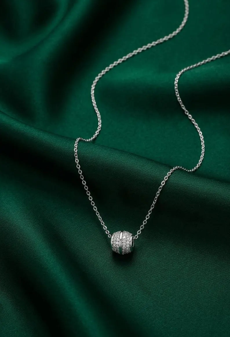 A delicate silver chain necklace with a small, round pavé-set pendant, displayed on draped emerald green satin fabric.