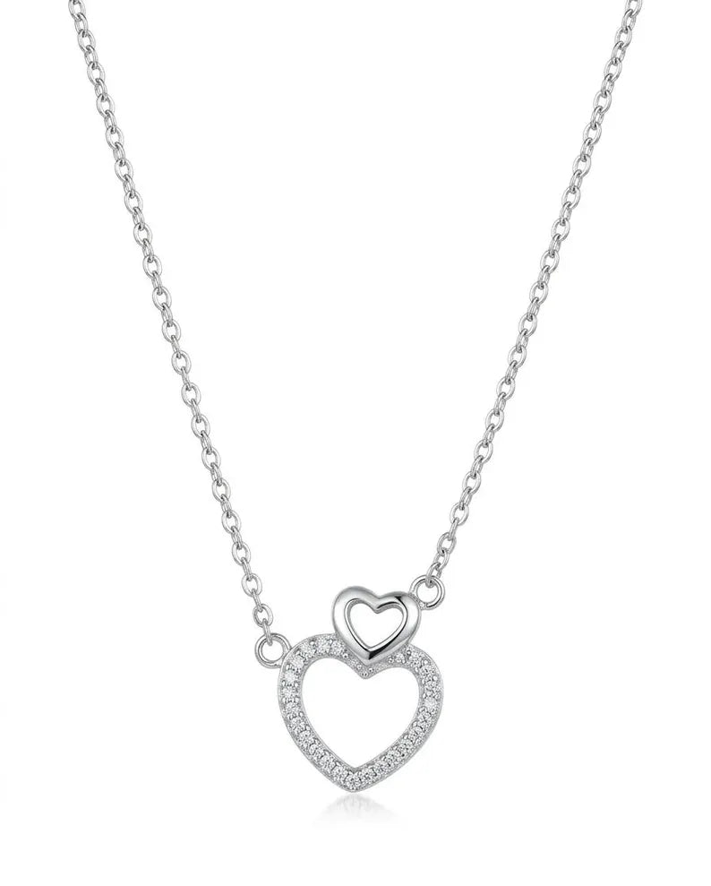 A silver heart pendant necklace featuring a smaller solid heart nested inside a larger open heart paved with small clear stones, suspended from a delicate silver chain on a white background.