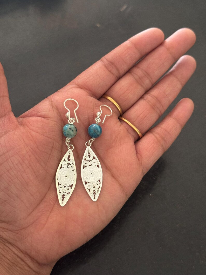 A pair of silver filigree dangle earrings with small turquoise-blue beads and silver fishhook wires, shown hanging from a person's open hand. The person is wearing two gold rings.