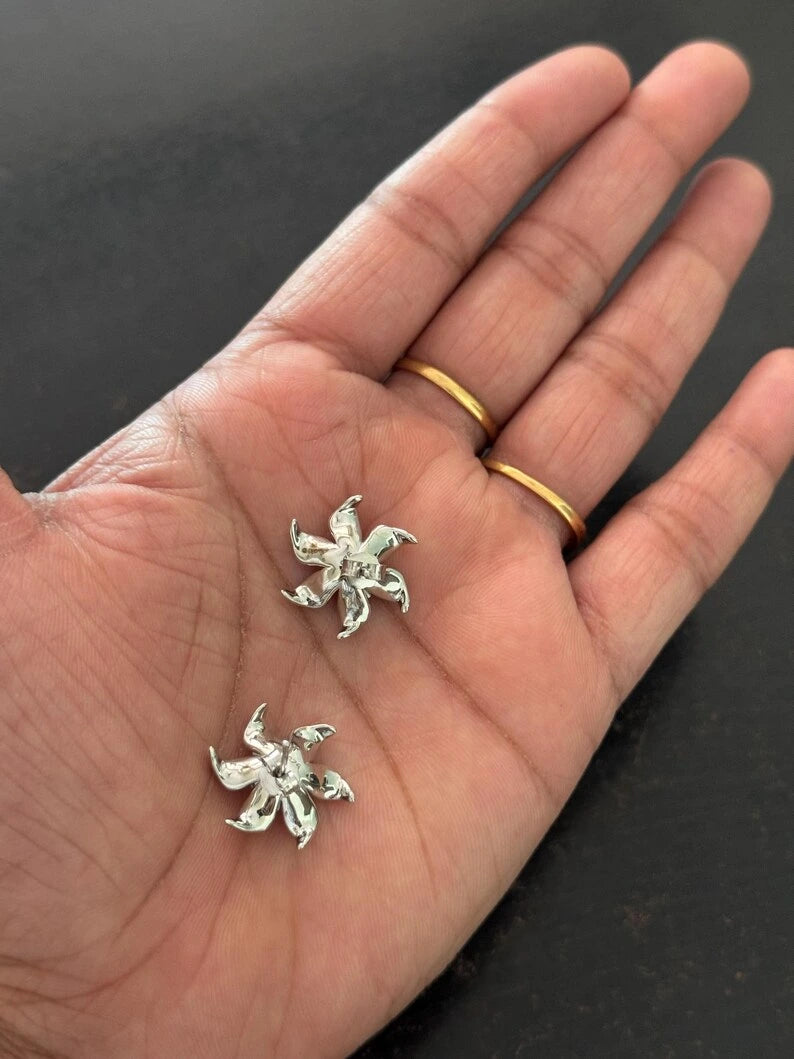A close-up of a person's open hand displaying a pair of small silver flower-shaped stud earrings, with two gold rings on the fingers, against a dark background.