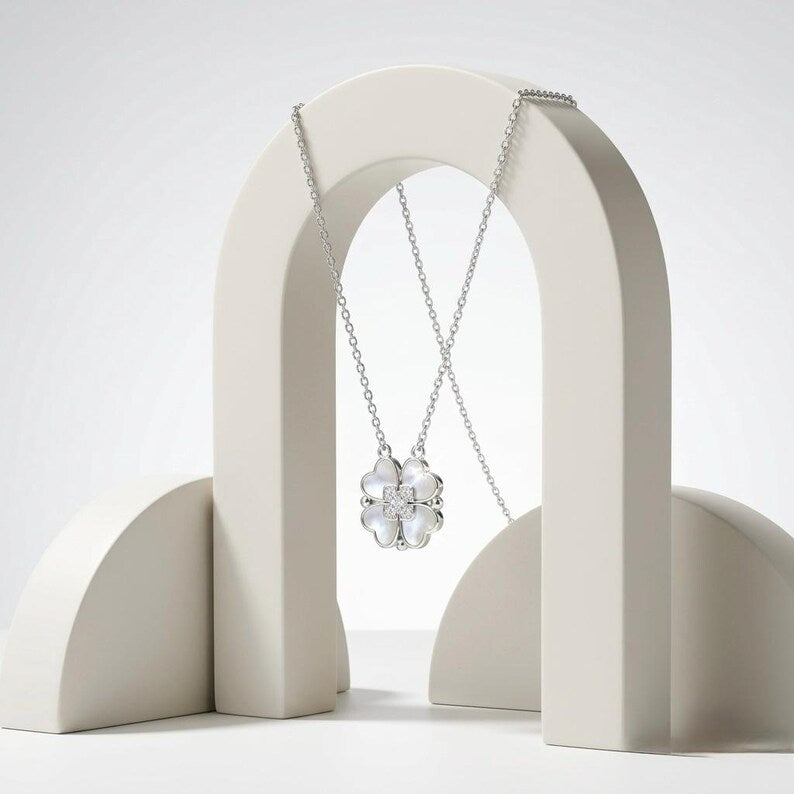 A silver-toned necklace with a clear faceted flower-shaped pendant, draped over a minimalist off-white arched display stand against a light gray studio background.