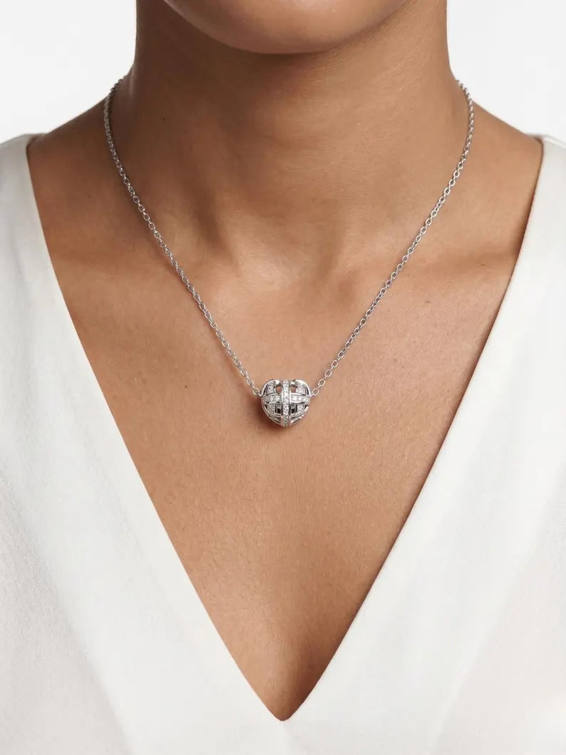 A close-up of a model wearing a silver chain necklace with a heart-shaped pendant featuring a cross design, styled with a white V-neck top.