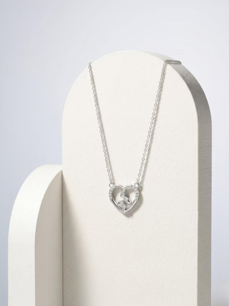 A silver heart pendant necklace with a central clear gemstone, displayed on a white arched jewelry stand against a light gray background.