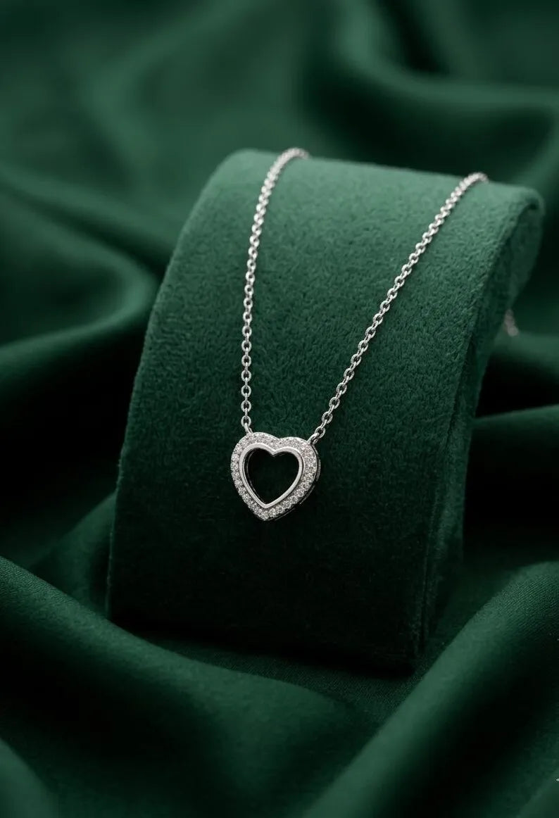 A silver heart pendant necklace with a small inlaid stone border, resting on a dark green velvet jewelry display stand against a draped dark green fabric background.