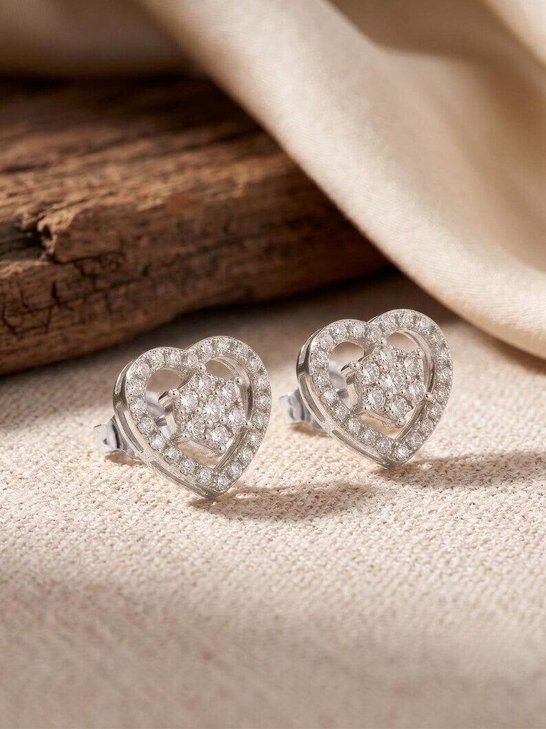 A pair of silver heart-shaped stud earrings set with clear pavé gemstones, displayed on a textured beige fabric next to a rustic piece of wood.