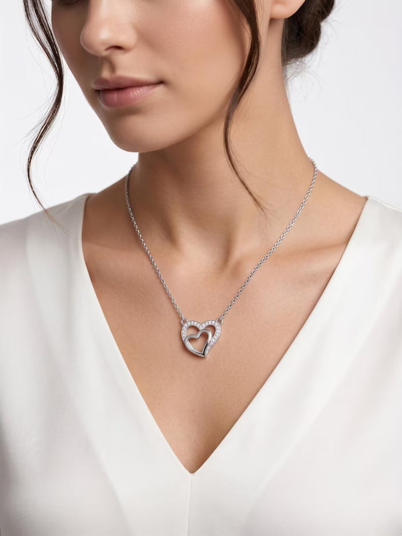 A close-up of a woman wearing a delicate silver heart-shaped pendant necklace with small clear stones, paired with a white V-neck top against a light gray background.