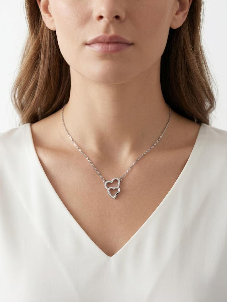 A close-up of a woman wearing a delicate silver necklace with a double-heart pendant, styled with a white V-neck top against a clean white background.
