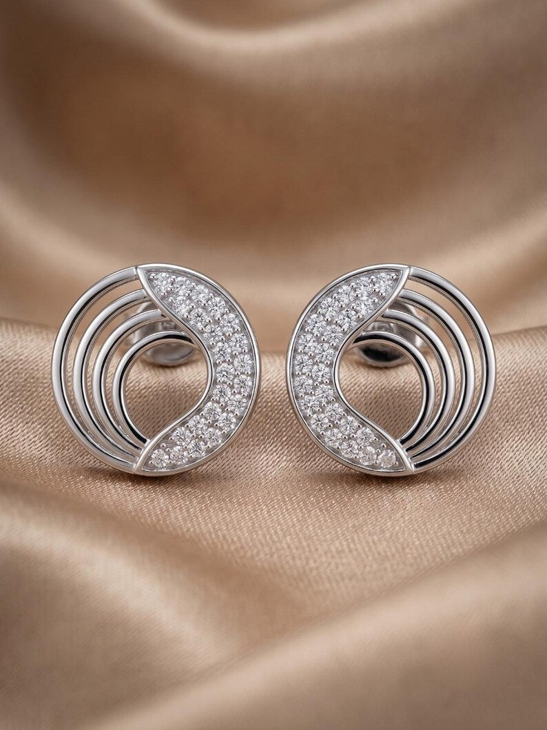 A pair of silver crescent-shaped stud earrings paved with clear stones, displayed on a draped beige satin fabric background.