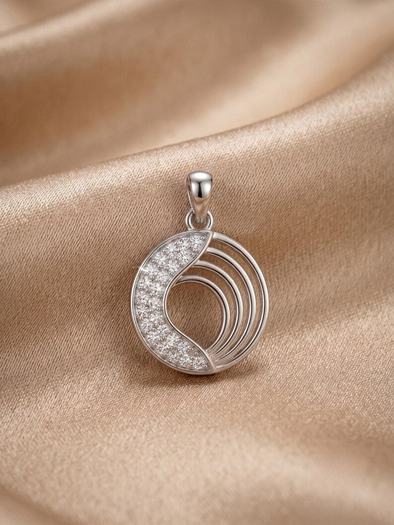 A silver crescent moon pendant with a pavé-set diamond half and concentric wire accents, displayed on draped champagne-colored satin fabric.