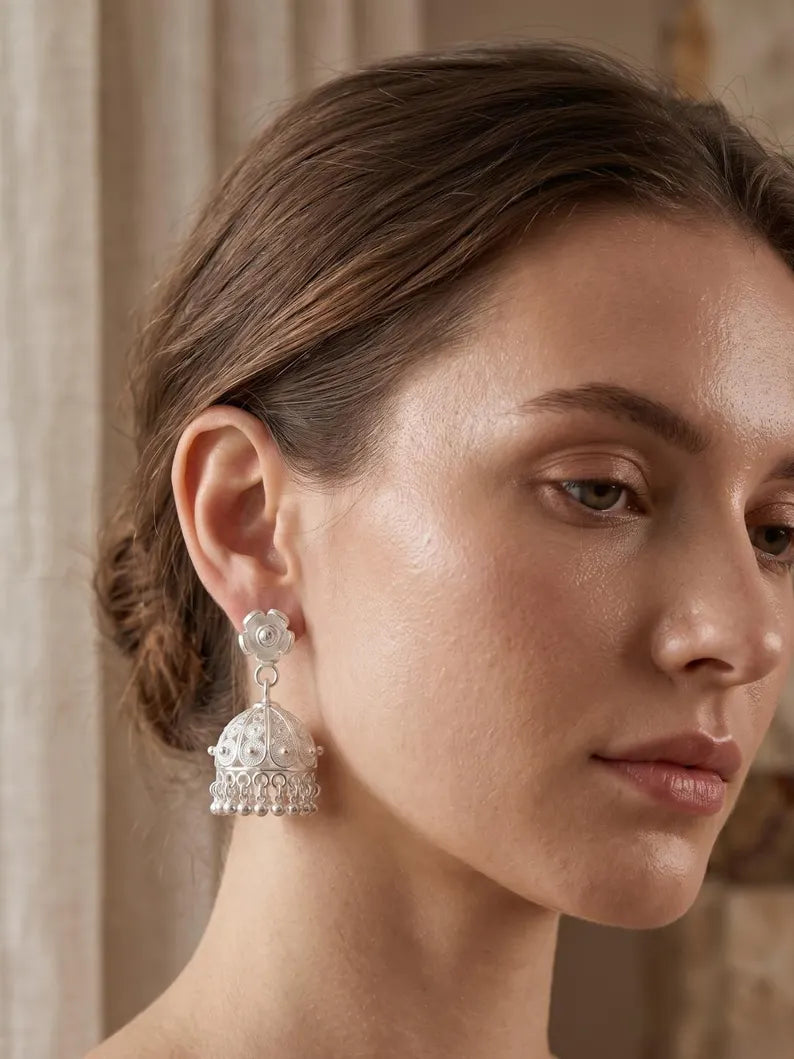A close-up of a woman wearing a silver-toned jhumka earring with a clear stone top and a dangling ornate bell-shaped charm, set against a soft beige background.