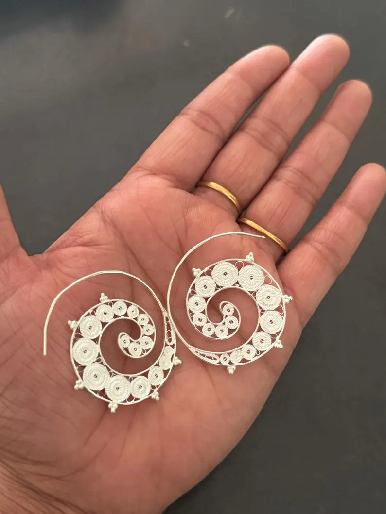A pair of large spiral-shaped silver earrings displayed on an open hand, with two gold rings worn on the fingers, against a dark gray background.