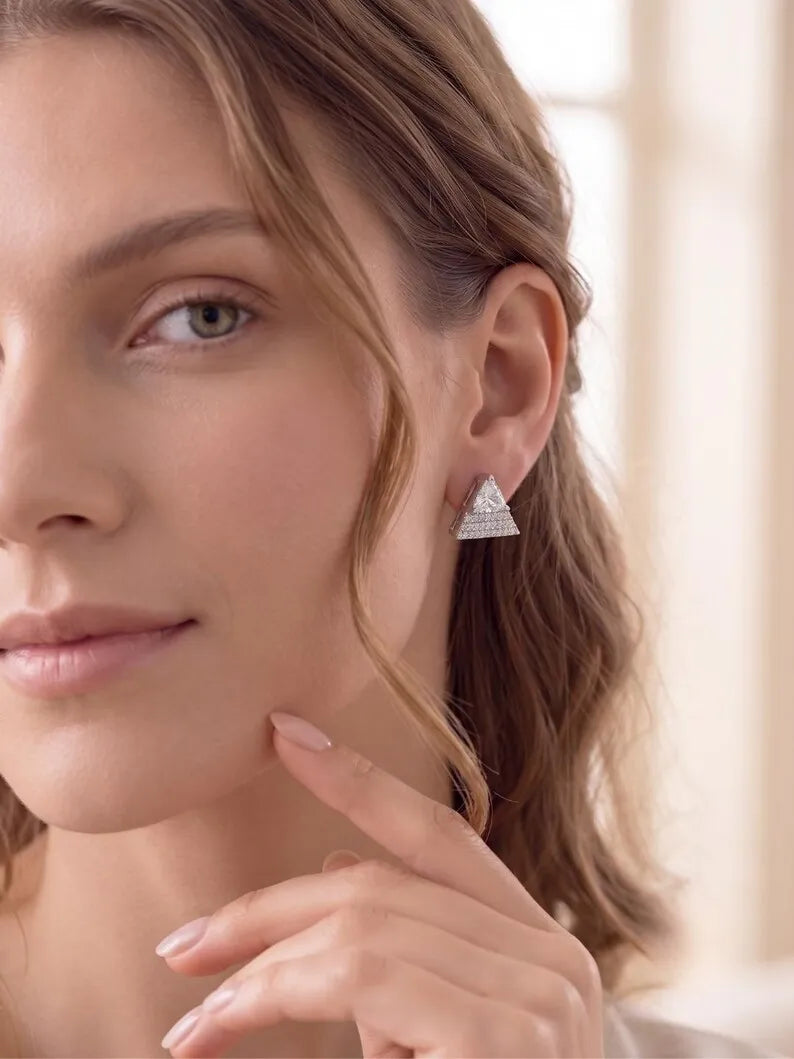 A close-up of a woman wearing a small triangular silver stud earring with a textured, diamond-like surface, set against a soft, warm-toned blurred background.