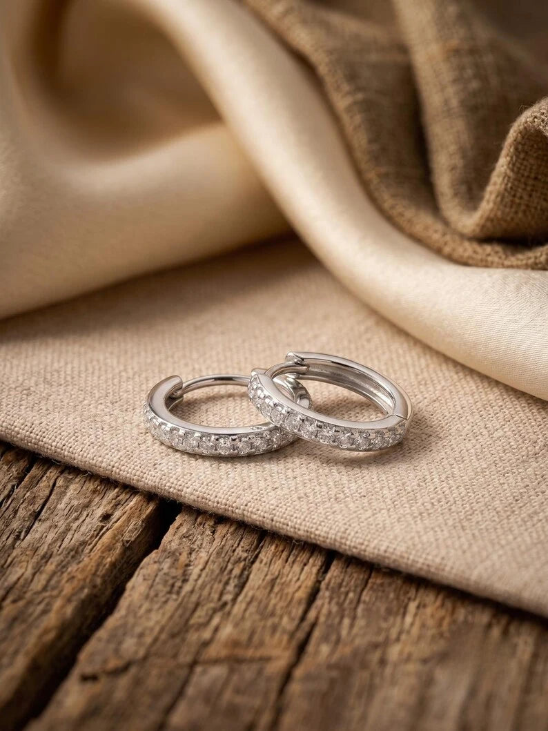 Two silver hoop rings set with small clear stones, resting on a beige fabric surface over a rustic wooden table.