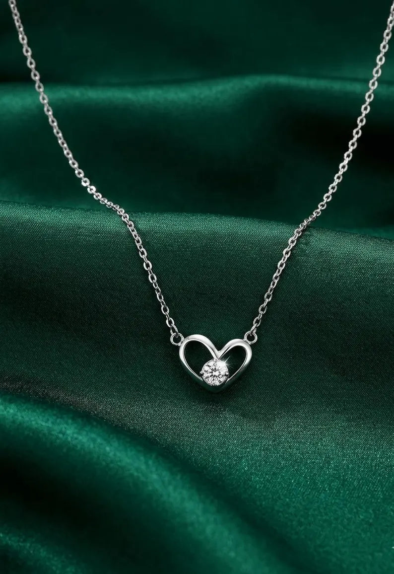 A silver heart pendant necklace with a clear round gemstone, displayed on a draped dark green satin fabric background.