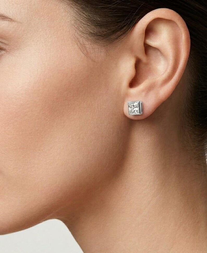 A close-up of a model's ear wearing a small square stud earring with a silver-toned metal border and clear faceted gemstone.