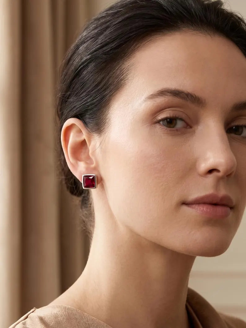 A close-up portrait of a woman wearing a small square stud earring with a deep red gemstone in a silver-toned bezel setting, against a soft beige background.