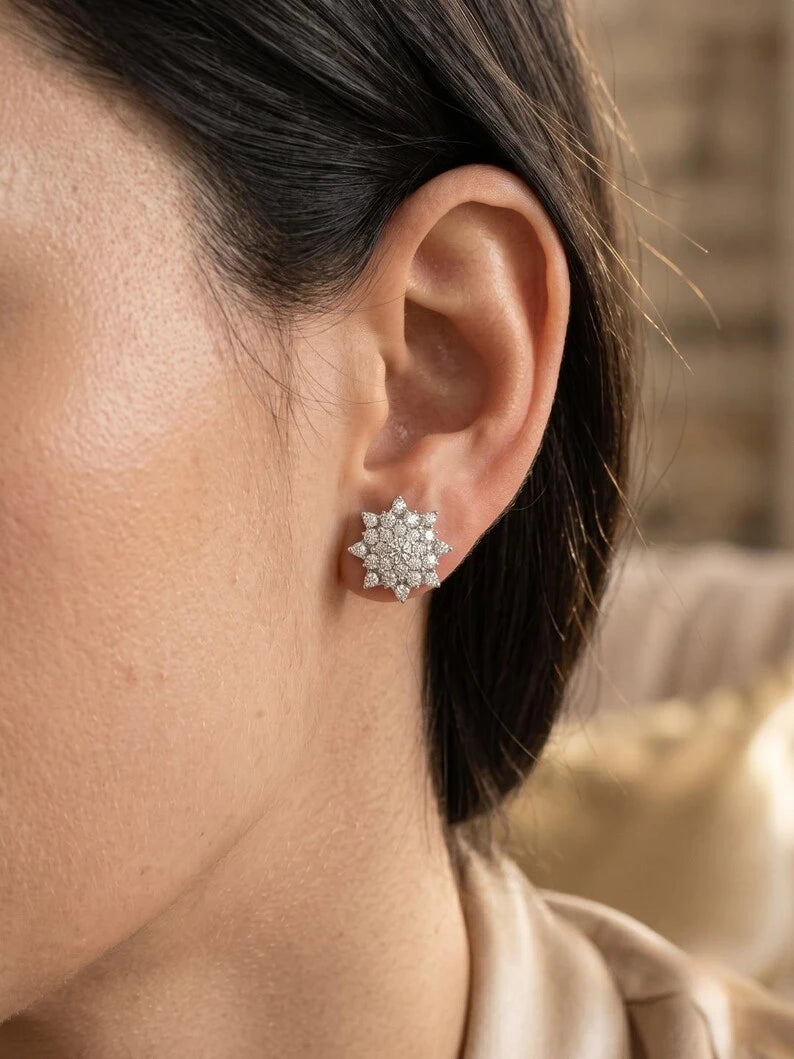 A close-up of a model's ear wearing a silver-toned starburst stud earring set with clear gemstones, against a softly blurred indoor background.
