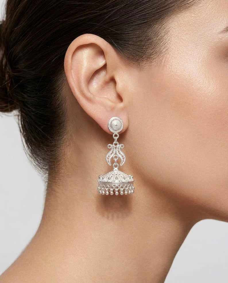 A close-up of a model's ear wearing a silver-toned traditional Indian jhumka earring with a round stud and a dangling ornate bell-shaped drop embellished with small clear stones.