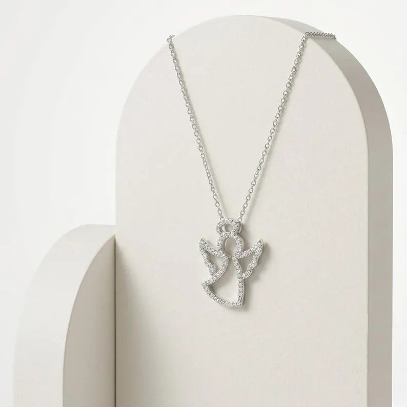 A silver-toned angel pendant necklace with small clear stones, displayed on a cream-colored arched jewelry stand against a soft white background.