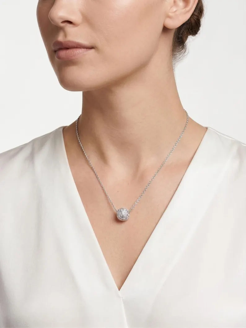 A close-up of a model wearing a delicate silver chain necklace with a round, clear pavé-set pendant, paired with a white satin V-neck top.