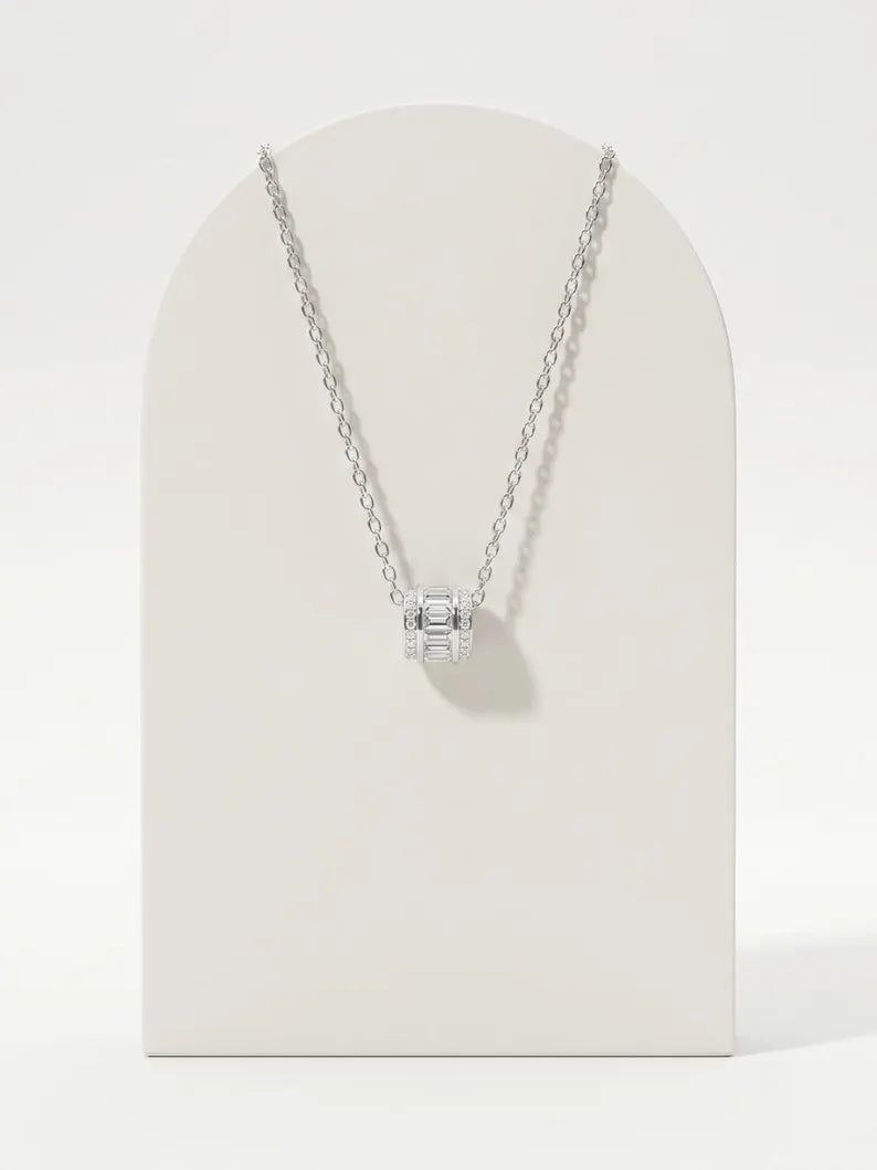 A silver chain necklace with a small box-shaped pendant set with clear stones, displayed on a cream-colored arched stand against a white background.