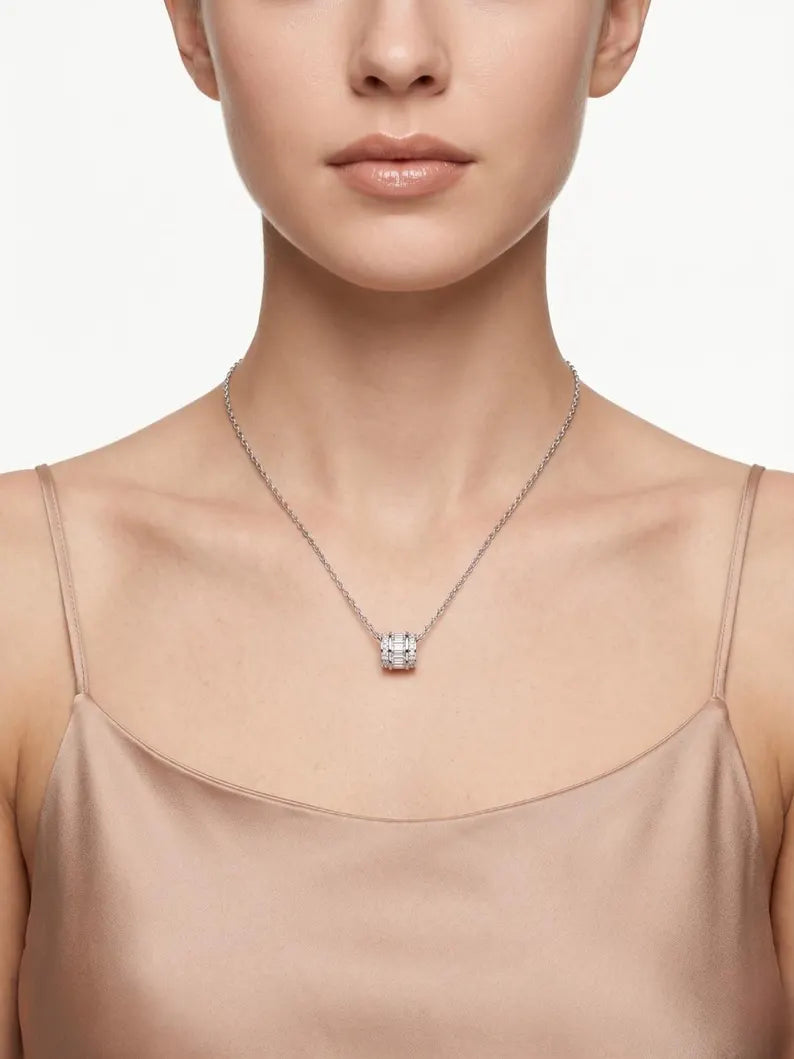 A close-up of a model wearing a delicate silver chain necklace with a small square pendant set with clear gemstones, paired with a beige satin camisole top against a clean white background.